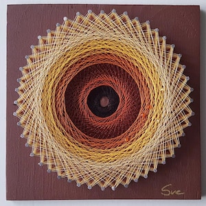 May include: A square dark brown wooden board features a circular string art design. The pattern uses yellow, orange, and dark red threads wrapped around silver nails to create a radial gradient effect. The signature "Sve" is written in gold.