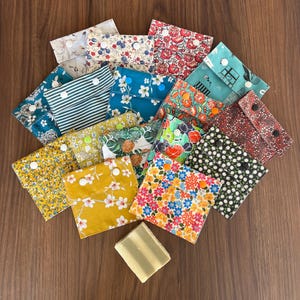 Könnte beinhalten: Eine Sammlung kleiner, quadratischer Stoffbeutel mit verschiedenen Blumen-, Streifen- und Mustermotiven in Farben wie Gelb, Blau, Rot, Grün und Schwarz. Jeder Beutel hat eine Klappe, die mit einem Kunststoff-Druckknopf verschlossen wird. Ein rechteckiges Stück hellgrüne Seife liegt unter den Beuteln.