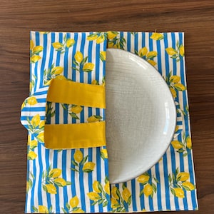 Può includere: Un piatto in ceramica bianca è posto all'interno di un supporto in tessuto a righe blu e bianche con un motivo a limoni. Il supporto ha cinghie gialle e una chiusura a linguetta gialla. Il tessuto è decorato con limoni gialli e foglie verdi.