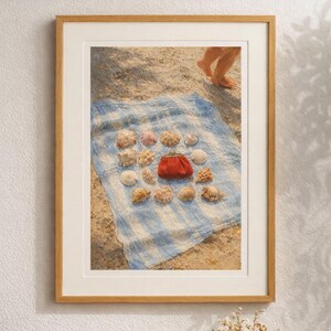 Könnte beinhalten: Ein gerahmter Druck mit einer Strandszene. Ein blau-weiß gestreiftes Handtuch auf dem Sand zeigt eine Sammlung von Muscheln und eine kleine rote Geldbörse. Das Bild ist in einem hellbraunen Holzrahmen gerahmt.
