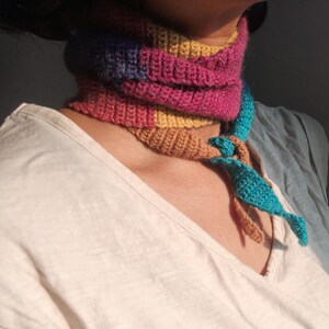 Op de afbeelding: Een kleurrijke, gebreide cowl sjaal in roze, geel, blauw en bruin. De sjaal is om de nek gewikkeld en heeft een turquoise accent met bruine kwastjes. De sjaal wordt gedragen over een wit t-shirt.