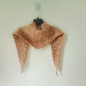 Könnte beinhalten: Ein gestrickter Schal in hellem Pfirsichton, der an einem schwarzen Kleiderbügel hängt. Der Schal hat eine dreieckige Form mit langen, spitzen Enden. Die Textur des Strickwerks ist sichtbar, und der Schal scheint aus weichem Garn zu bestehen.