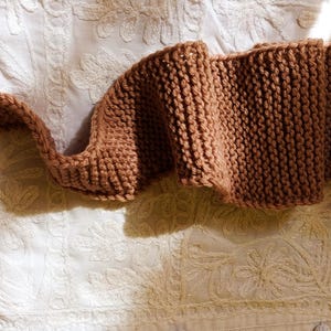 Op de afbeelding: Een bruine gebreide sjaal, weergegeven in een golvend patroon. De sjaal is gemaakt van dik garen, met een zichtbare breistructuur. De sjaal ligt op een wit, getextureerd oppervlak.