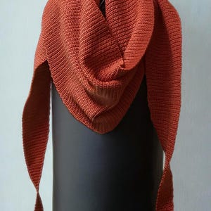 Può includere: Uno scialle triangolare color ruggine, realizzato in tessuto a maglia. Lo scialle è drappeggiato su un oggetto cilindrico nero e sospeso da una catena di metallo. Le estremità dello scialle hanno un design a punta.