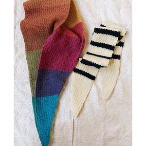 Op de afbeelding: Een handgebreide sjaal en beenwarmers. De sjaal heeft een kleurovergang van bruin, groen, paars, blauw en turquoise. De beenwarmers zijn crèmekleurig met zwarte strepen.