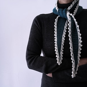 以下が含まれることがあります： 人々の首にドレープされた、スカラップエッジのティールと白のかぎ針編みのスカーフ。スカーフは、白い装飾的なトリムが付いた濃いティール糸で作られています。その人は濃い灰色のセーターを着ています。