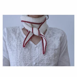 May include: A white knitted scarf with a red trim is tied around the neck. The scarf is made of a soft, textured yarn. The red trim adds a pop of color to the otherwise neutral scarf. The scarf is a stylish accessory.