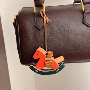 Puede incluir: Bolso de mano de cuero marrón oscuro con detalles dorados y un pequeño adorno de caballo balancín de madera. El adorno es naranja, verde y marrón. El bolso tiene un asa superior y cierre de cremallera.