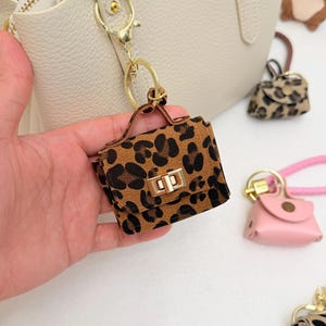Può includere: Un portachiavi a forma di mini borsa con stampa leopardata, con chiusura e manico color oro. Il portachiavi è attaccato a una borsa bianca con cerniera dorata. Altri piccoli portachiavi a forma di borsa sono visibili sullo sfondo.