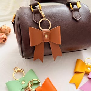 Puede incluir: Un llavero de lazo de cuero marrón sujeto a un bolso marrón. Se ven llaveros adicionales en verde, naranja, amarillo y rosa. Los llaveros de lazo tienen herrajes dorados.