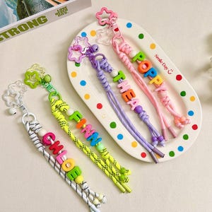 May include: Four braided cord keychains with star-shaped clasps and colorful letter beads spelling the names Chloe, Finn, Thea, and Poppy. The keychains feature white, neon yellow, purple, and pink cords with matching beads and small bells.