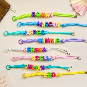 May include: Eight colorful cord bracelets, each featuring a unique name spelled out in plastic letter beads. The bracelets include names such as Rowan, Marcus, Jasper, Winnie, Serena, Stella, Julian, and Leo, with metal end caps on the cords.