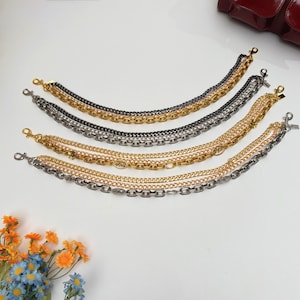 Puede incluir: Cuatro correas de cadena en capas en tonos dorados, plateados y negros. Cada correa presenta un diseño de eslabones de cadena diferente e incluye un cierre en cada extremo para una fácil fijación a una bolsa. Las cadenas están dispuestas sobre una superficie blanca.