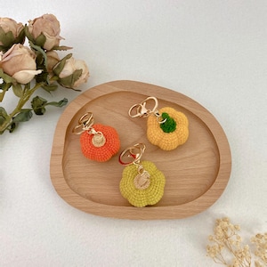 Puede incluir: Tres llaveros de calabaza tejidos a ganchillo en naranja, amarillo y verde claro, cada uno con una anilla de metal dorado y un pequeño detalle de tallo beige o verde. Las calabazas tienen textura de hilo y una forma redondeada y segmentada.