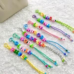 Puede incluir: Llaveros de cuentas de colores que deletrean nombres como "Lara", "Anna" y "Aurora". Cada llavero presenta una combinación de colores y cordón diferente, con un cierre de metal. Las cuentas son de varios colores brillantes.