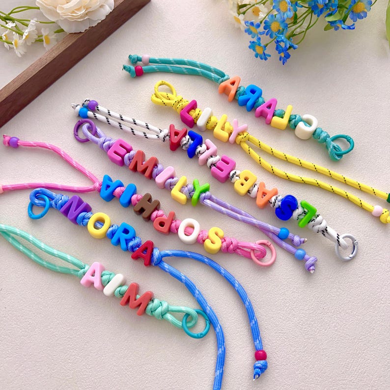 Peut inclure: Plusieurs bracelets de noms color&eacute;s. Chaque bracelet est compos&eacute; de perles de lettres vives &eacute;pelant un nom diff&eacute;rent. Les cordons sont de diff&eacute;rentes couleurs, notamment le bleu, le jaune et le rose. Les bracelets sont pr&eacute;sent&eacute;s sur une surface claire.