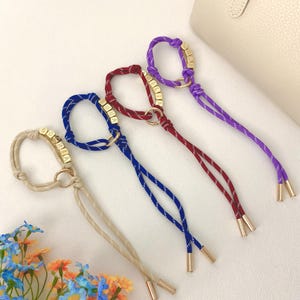 Könnte beinhalten: Vier farbige Kordelarmbänder mit goldfarbener Hardware und Namensperlen. Die Armbänder sind in Beige, Blau, Rot und Lila gehalten. Jedes Armband hat eine Schlaufe und zwei herabhängende Kordeln mit goldfarbenen Spitzen. Das Wort "SOPHIA" ist auf dem beigen Armband geschrieben.