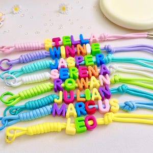 Puede incluir: Una colección de cordones coloridos con cuentas de letras de plástico que forman los nombres Stella, Winnie, Jasper, Serena, Rowan, Julian, Marcus y Leo. Cada cordón está hecho de cuerda trenzada en varios colores pastel y brillantes.