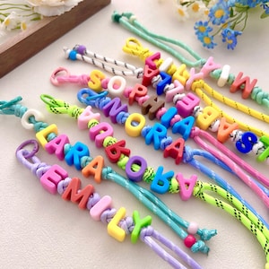 Puede incluir: Pulseras de cuentas coloridas que deletrean nombres como "Emily", "Lara" y "Sophia". Cada pulsera presenta cuentas de letras vibrantes ensartadas en un cordón con una variedad de colores, incluyendo rosa, azul, amarillo y verde. Un accesorio divertido y personalizado.