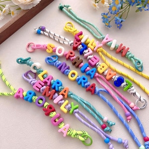 Peut inclure: Bracelets de noms color&eacute;s avec des noms &eacute;pel&eacute;s en lettres majuscules vives. Les bracelets pr&eacute;sentent une vari&eacute;t&eacute; de couleurs de cordon, notamment rose, bleu, jaune et vert. Chaque bracelet est unique, avec diff&eacute;rentes combinaisons de couleurs et de noms.