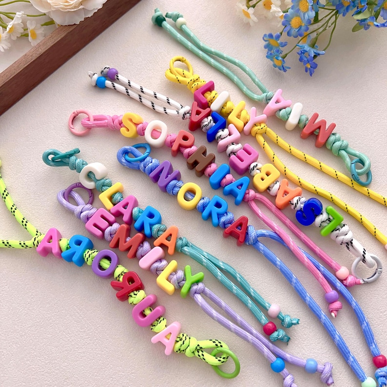 Peut inclure: Bracelets de noms color&eacute;s avec des noms &eacute;pel&eacute;s en lettres majuscules vives. Les bracelets pr&eacute;sentent une vari&eacute;t&eacute; de couleurs de cordon, notamment rose, bleu, jaune et vert. Chaque bracelet est unique, avec diff&eacute;rentes combinaisons de couleurs et de noms.