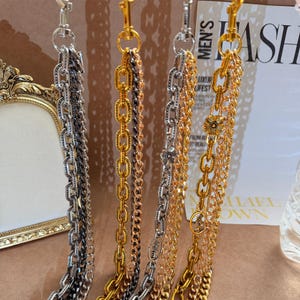 Puede incluir: Primer plano de varias correas de cadena decorativas en tonos dorados, plateados y bronce. Cada cadena tiene un cierre en la parte superior. Algunas cadenas tienen dijes florales decorativos. El fondo incluye una revista con la palabra "MEN'S".