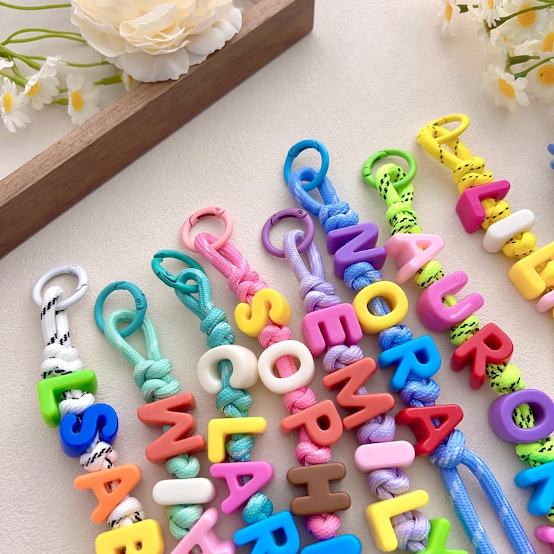 Peut inclure: Plusieurs porte-cl&eacute;s color&eacute;s avec des perles de lettres &eacute;pelant des noms. Chaque porte-cl&eacute;s pr&eacute;sente une palette de couleurs diff&eacute;rente, avec des lettres de couleurs vives comme le rose, le jaune, le bleu et le vert. Les porte-cl&eacute;s sont attach&eacute;s &agrave; un mousqueton en m&eacute;tal.