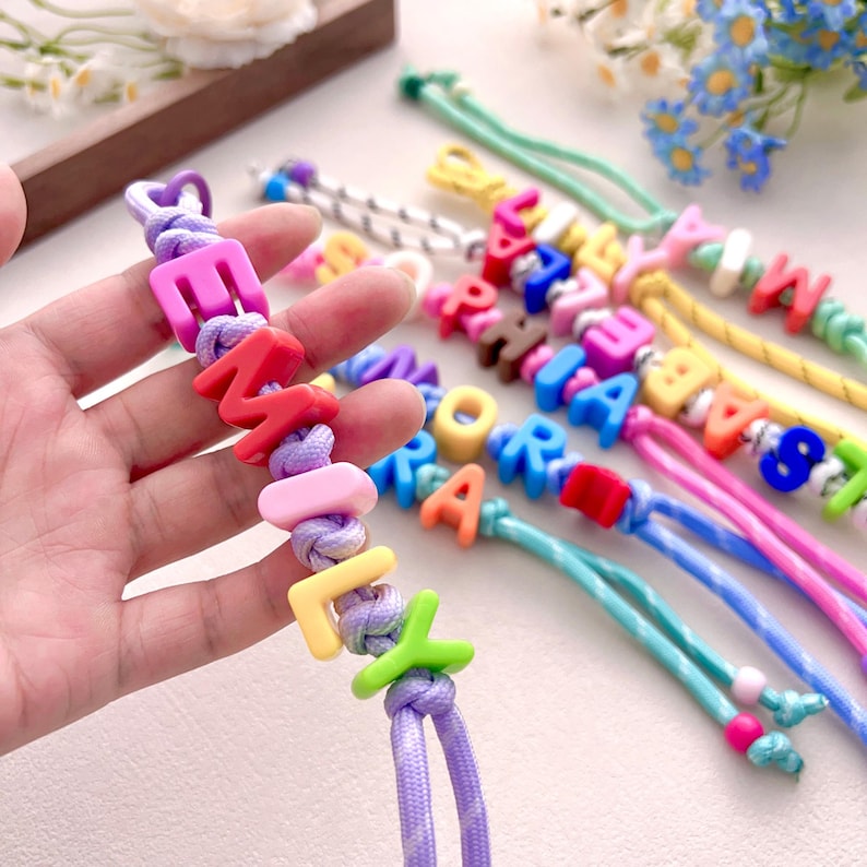 Peut inclure: Gros plan sur des bracelets de perles color&eacute;es &eacute;pelant des noms. Les bracelets pr&eacute;sentent diff&eacute;rentes couleurs de perles et de cordons. Les lettres sont de diff&eacute;rentes couleurs, dont le rose, le rouge, le jaune et le vert. Les bracelets sont sur une surface blanche.