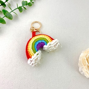 May include: A colorful crochet rainbow keychain with a gold-colored ring. The rainbow features red, orange, yellow, green, blue, and purple stripes. White fluffy clouds are attached to the bottom of the rainbow.