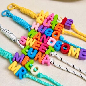 Può includere: Quattro cordini in nylon colorato, ciascuno con perline a lettera in plastica che compongono le parole MAISIE, CUTE, HUGO, HANDSOME, SWEET, LUNA, MILO e MR. I cordini sono gialli, blu e bianchi con macchioline nere.