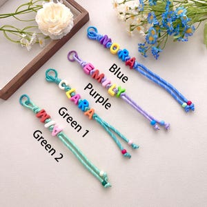 Peut inclure: Quatre porte-cl&eacute;s personnalis&eacute;s faits &agrave; la main, bleus, violets et verts. Chaque porte-cl&eacute;s pr&eacute;sente un nom &eacute;pel&eacute; avec des perles de lettres color&eacute;es. Les noms sont "Noora", "Emily", "Clara" et "Mia".