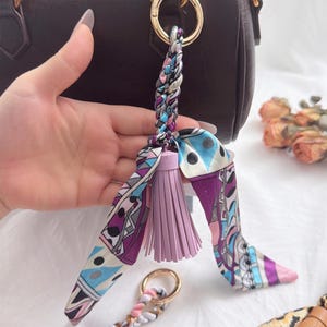 Puede incluir: Un charm decorativo para bolso con un cordón trenzado, una borla lavanda y un pañuelo de seda estampado en tonos morados, azules y rosas. El charm está sujeto a un anillo dorado, diseñado para complementar un bolso.
