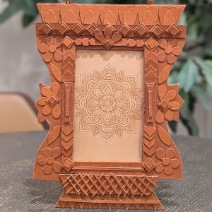 May include: A decorative wooden picture frame with a detailed floral and geometric design. The frame is a warm brown colour and features a central panel with a mandala design. The frame is approximately 25 cm tall.