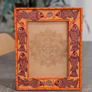 May include: A rectangular orange picture frame featuring hand-painted fish motifs along the border. The frame displays six fish in shades of brown and red, accented with small white dots. Inside the frame is a tan insert with a brown mandala design.