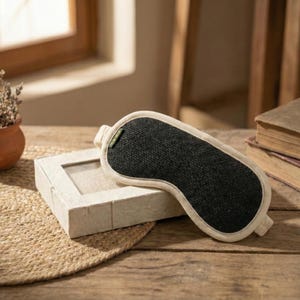 May include: A black sleep mask with a cream-colored border rests on a wooden surface next to a small, rectangular box. A small terracotta pot with dried plants and a stack of books are also visible. The mask is designed to block out light.