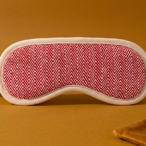 May include: A sleep mask with a red and white herringbone pattern. The mask has a cream-coloured border and matching elastic strap. The mask is designed to block out light for better sleep. A small, brown pouch is in the background.