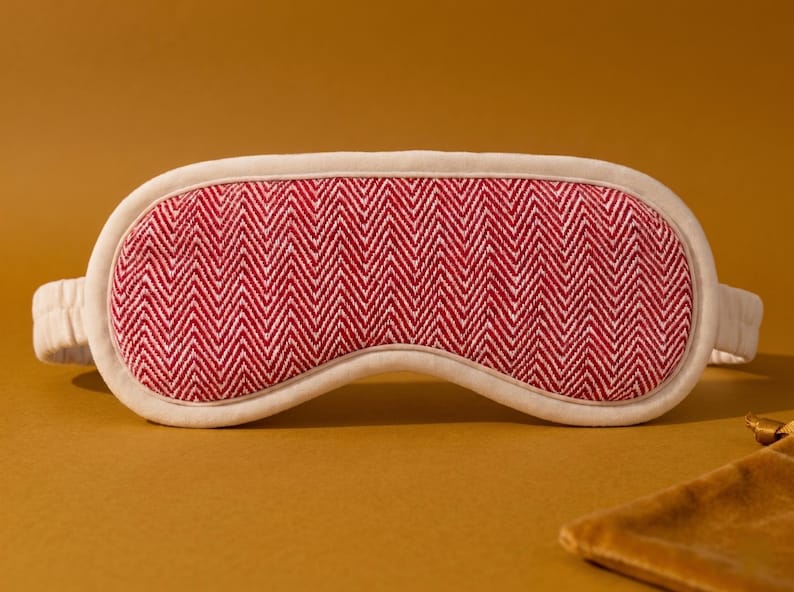 May include: A sleep mask with a red and white herringbone pattern. The mask has a cream-coloured border and matching elastic strap. The mask is designed to block out light for better sleep. A small, brown pouch is in the background.