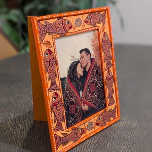 May include: A rectangular orange picture frame featuring hand-painted fish motifs in shades of brown and red along the border. The frame contains a photograph of two people wrapped in a patterned blanket, with white dot accents along the edges.