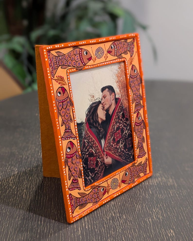 May include: A rectangular orange picture frame featuring hand-painted fish motifs in shades of brown and red along the border. The frame contains a photograph of two people wrapped in a patterned blanket, with white dot accents along the edges.