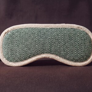 May include: A sleep mask featuring a green and white herringbone design. The mask has a white trim and elasticated straps. It is designed to block out light for improved sleep. The mask is made from soft fabric.