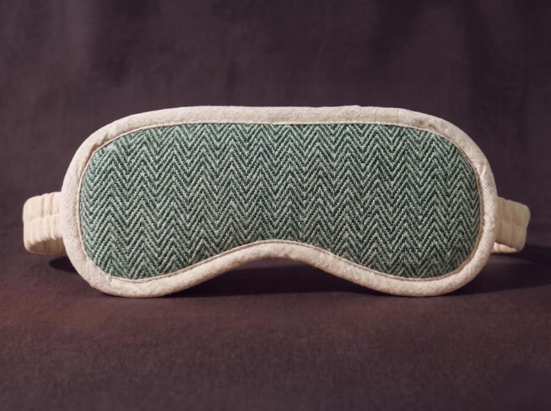 May include: A sleep mask featuring a green and white herringbone design. The mask has a white trim and elasticated straps. It is designed to block out light for improved sleep. The mask is made from soft fabric.