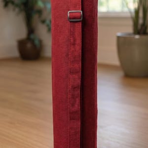 May include: A cylindrical dark red fabric carrying bag with a textured weave. The bag features a long, adjustable vertical strap with a dark plastic buckle and a drawstring closure at the top with a black cord lock for securing the contents inside.