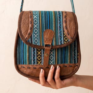 May include: A brown leather crossbody bag with a flap closure. The bag features a colorful woven fabric panel with blue, green, yellow, and black stripes and geometric patterns. The bag is held by a hand.