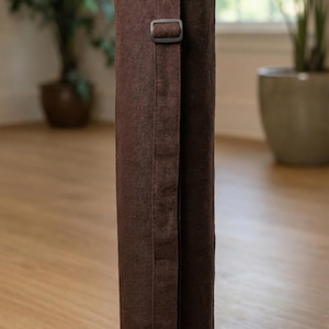 May include: A cylindrical dark brown yoga mat bag made of a textured fabric. The bag features a long, vertical strap running down the center with a black plastic adjustable buckle for carrying. The top of the bag is closed with a circular fabric lid.