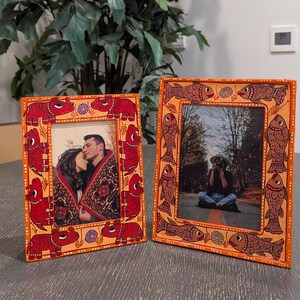 May include: Two rectangular picture frames with orange borders. The left frame features hand-painted red rhinoceros figures and small purple flowers. The right frame displays hand-painted brown fish figures and small purple flowers. Both frames hold photos.