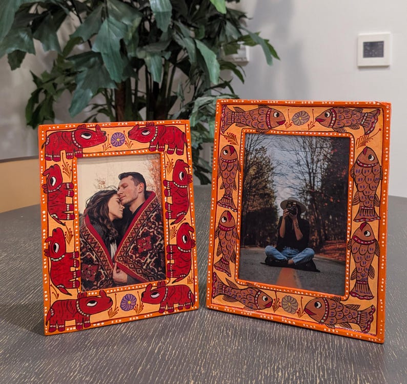 May include: Two rectangular picture frames with orange borders. The left frame features hand-painted red rhinoceros figures and small purple flowers. The right frame displays hand-painted brown fish figures and small purple flowers. Both frames hold photos.