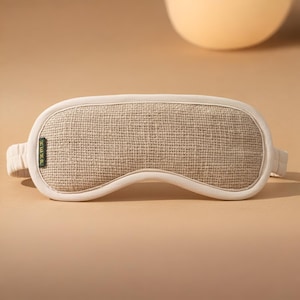May include: A beige sleep mask with a woven texture and a cream-colored border. The mask has a band on each side for securing it around the head. The word "HEMP" is printed on a small green tag on the left side of the mask.