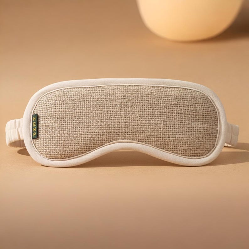 May include: A beige sleep mask with a woven texture and a cream-colored border. The mask has a band on each side for securing it around the head. The word "HEMP" is printed vertically on a small green tag on the mask.