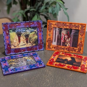 May include: Four rectangular picture frames with hand-painted designs. Two frames are purple with blue and red fish motifs, while two are orange with brown and red fish patterns. Each frame holds a different photograph, displaying various scenes.
