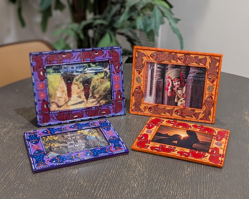 May include: Four rectangular picture frames with hand-painted designs. Two frames are purple with blue and red fish motifs, while two are orange with brown and red fish patterns. Each frame holds a different photograph, displaying various scenes.